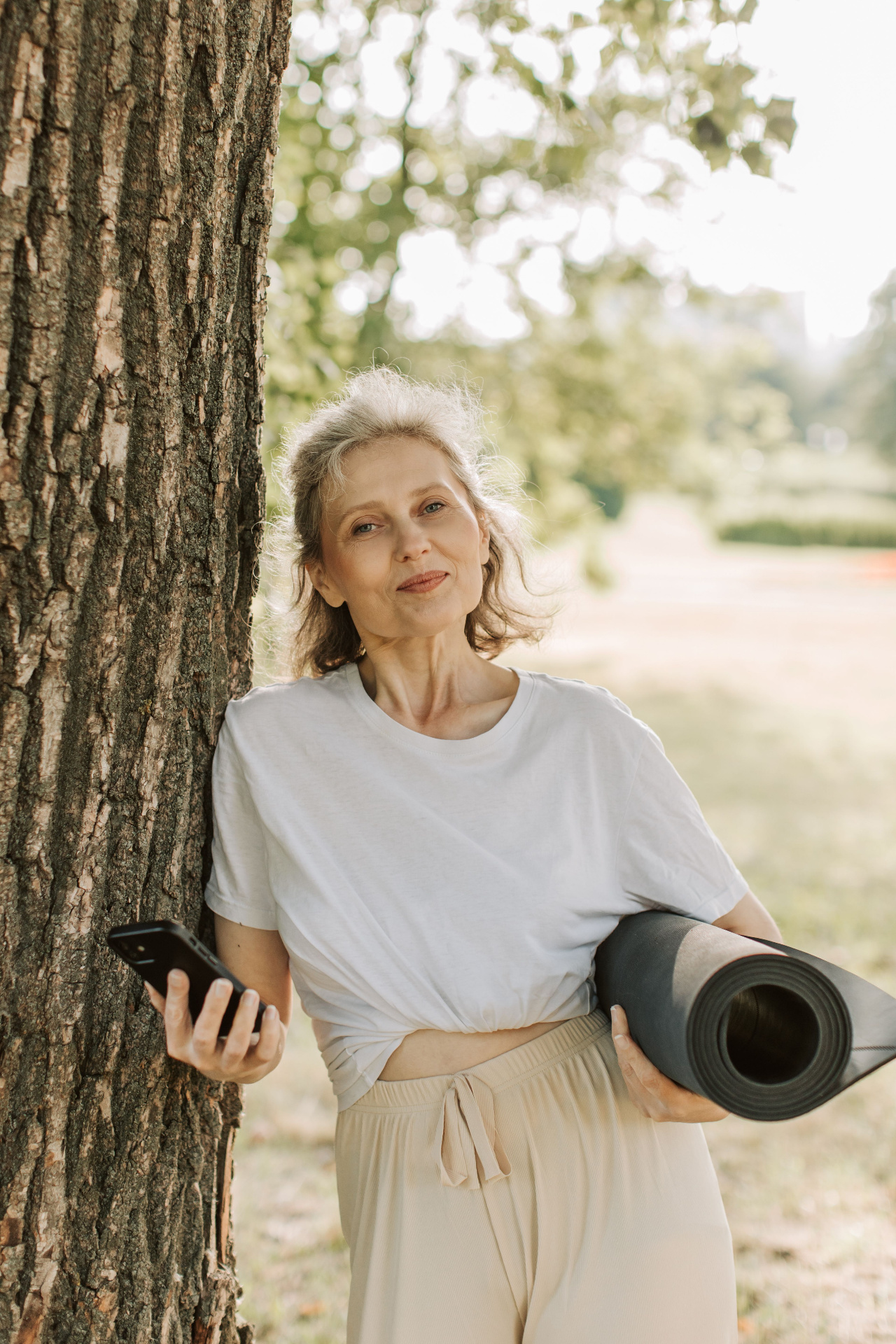 Mature woman outdoors with a yoga mat, leaning against a tree